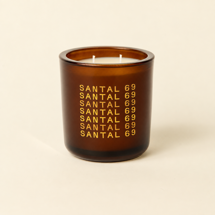 An amber glass jar candle with a white wax fill and gold text reading 'SANTAL 69' repeated vertically, set against a solid cream background.